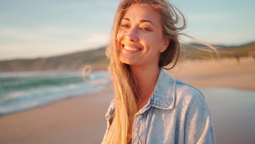Woman wearing denim shirt standing near sea in gentle breeze. Smiling long-haired lady enjoying evening walk by sea. Blonde woman in evening rays of sun at beach. Medium shot
