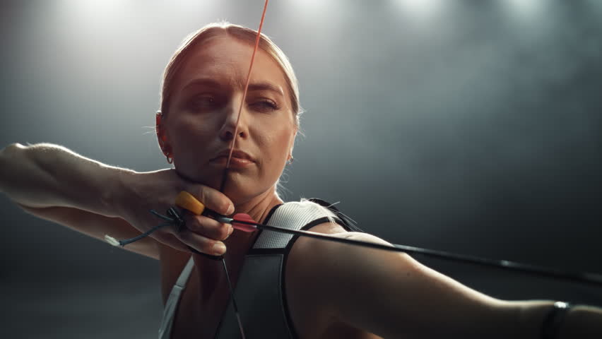 Slow Motion Speed Ramp Portrait Footage of a Female Archer Aiming and Shooting at a Target with a Modern Compound Bow. Young Athlete Drawing and Releasing the Arrow in a Dark Studio for Advertising Royalty-Free Stock Footage #1110269869