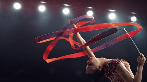 Portrait of a Graceful Rhythmic Gymnast Performing a Dance with a Red Ribbon. Young Female Exercising in a Striking Beautiful Uniform in a Dark Studio. Cinematic Super Slow Motion Speed Ramp Footage Adlı Stok Video
