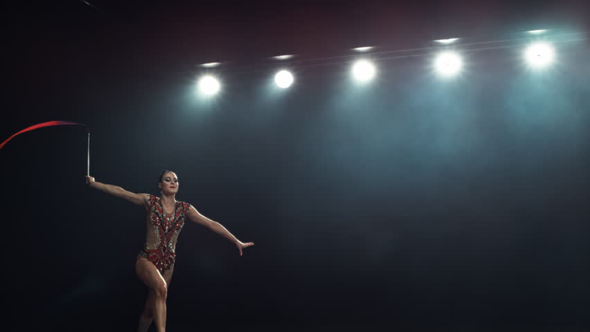 Graceful Rhythmic Gymnast Performs a Powerful Split Leap with a Ribbon. Young Female Exercising in a Striking Beautiful Uniform in a Dark Room. Cinematic Super Slow Motion Speed Ramp Footage