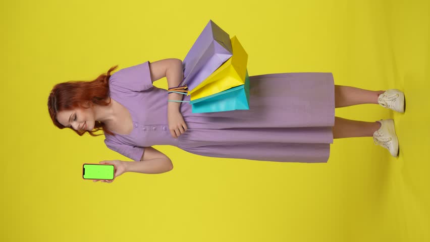 An attractive woman with shopping bags points her index finger at the green screen of her smartphone and shows a thumbs up. Young woman full length isolated on yellow. Vertical video. Advertising area Royalty-Free Stock Footage #1110272535