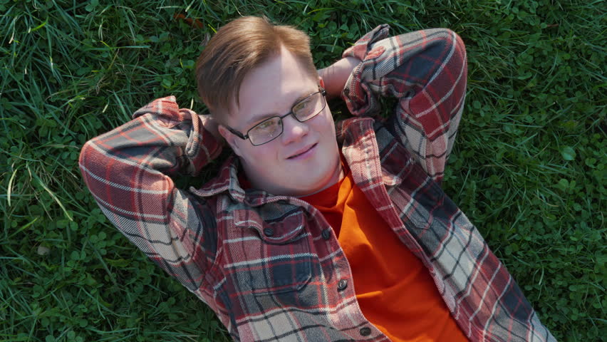 Portrait of young man with down syndrome in checkered shirt laying on the grass outdoors in the public park. Man with trisomy 21 illness. Disabled guy having a walk. Royalty-Free Stock Footage #1110280027
