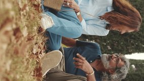 Vertical video, Retired couple talking, sitting on the grass in the park in autumn
