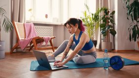 Young Asian woman with tattoos in sportswear exercising at home in the living room on a rubber mat with a laptop on video lessons. Sports, yoga and active lifestyle.