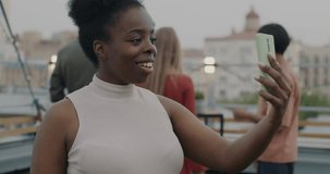 Happy African American woman making online video call with smartphone pointing at dancing people at rooftop party. Communication and modern devices concept.