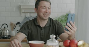 Middle aged man making online video call with smartphone talking gesturing sitting at kitchen table at home. Internet communication and modern lifestyle concept.