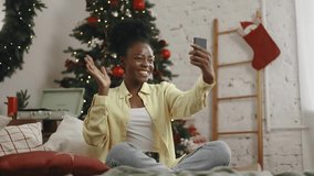 Beautiful young woman have video call conference meeting conversation on smartphone sitting on bed near Christmas tree with decoration at cozy home Happy smiling lady talking by smartphone indoors