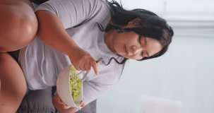 Vertical clip. A chubby Asian woman enjoy eating salad while relaxing on the sofa at home.