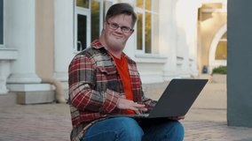 Young man with down syndrome in checkered shirt using laptop working online outdoors in the city. Guy with disability using laptop to make remote video call.