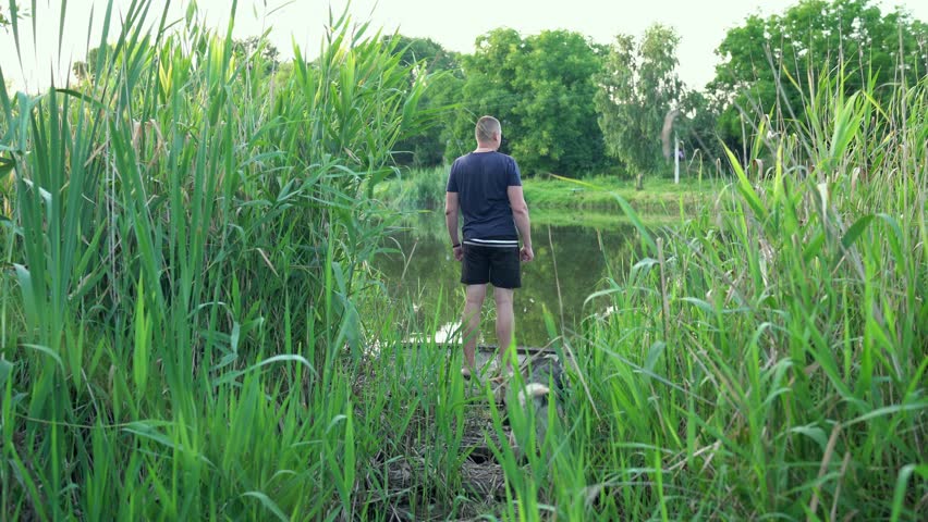 Young man with dog standing on the end of a jetty among the reeds and admires nature. Lonely man on a wooden pier looking into the distance in summer. People and pets. Nature video footage in 4K 25FPS