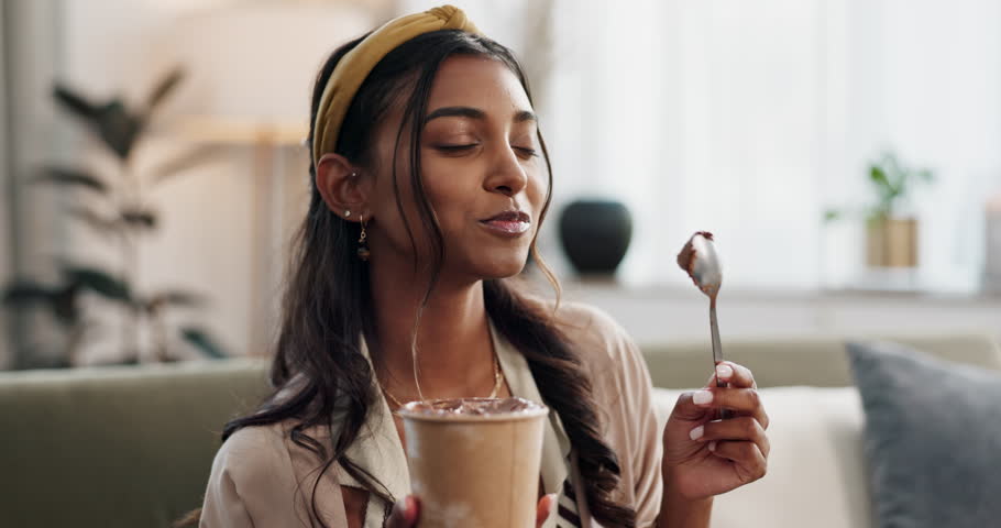 Woman, eating chocolate ice cream and sofa with happiness, gen z aesthetic and relax weekend in living room. Young, indian person and smile by dessert in excited, sweet tooth and sugar candy at home