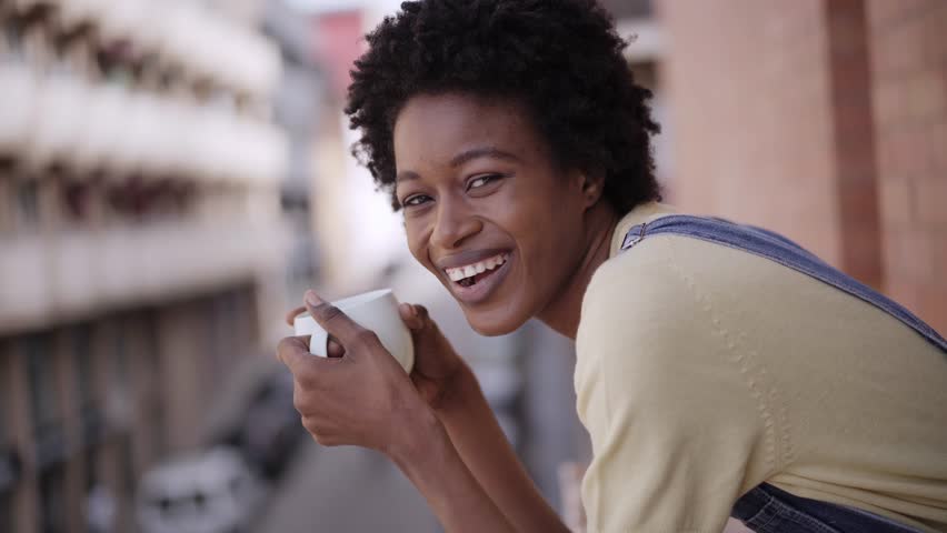 Cheerful African American woman poses looking at smiling camera leaning on floor balcony railing. Young black afro female drinking hot beverage and holding cup coffee outdoors. Domestic life at home.