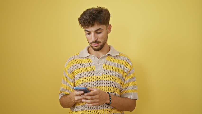 Upbeat young arabian man celebrating his win, confidently talking on smartphone, standing with a big smile against a yellow isolated background
