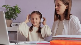 Brown haired little girl with pigtails sitting in front of laptop with her mother woman help with homework child wearing headphones being ready to start online lesson.