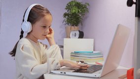 Online lesson. Distant education. little girl with pigtails wearing white shirt sitting at desk in front laptop showing greeting gesture for her teacher.