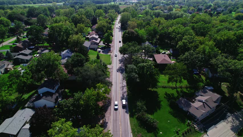 Aerial drone shot follows a straight road surrounded by American houses adorned with lush green gardens in an American neighborhood. Cars gently drive through the road in Crystal Lake, Illinois.