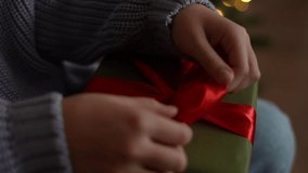 Close-up hands of unrecognizable little girl holding and using red ribbon to tie bow for wrapped gift box for Christmas present, on blurred background bright bokeh lights of Xmas tree, slow motion. - Powered by Shutterstock - Get 15% off with code: PIKWIZARD15