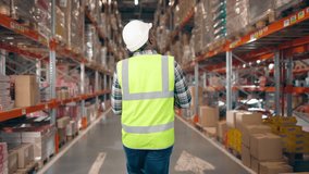 Back view of African American man walking at warehouse. Man in reflective vest and protective helmet. Male storage worker going at background of multi tiered racks. Distribution company. - Powered by Shutterstock - Get 15% off with code: PIKWIZARD15