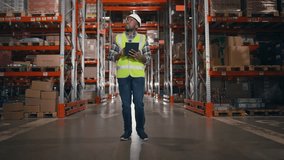 Contractor walking along storage of boxes with goods. African American man in helmet and reflective jacket. Inspector expertising warehouse stock. Male holding tablet and making notes. - Powered by Shutterstock - Get 15% off with code: PIKWIZARD15