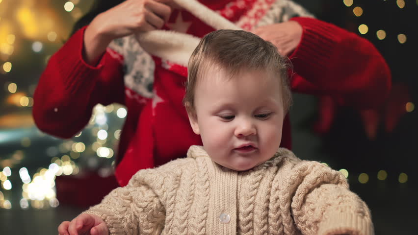 Mom put on sweet little funny baby boy red Santa hat at background of decorated fir-tree celebrate Christmas together. Happy family at holidays home. Curious child portrait. Merry Christmas, new year Royalty-Free Stock Footage #1110348467