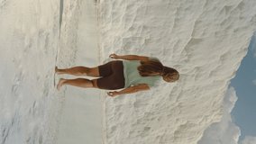 Vertical video. A young woman walks alone in the water on the white travertines of Pamukkale, with a backdrop of white walls adorned with calcite stalactites.