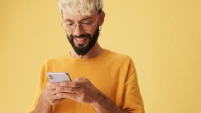 Guy with glasses, dressed in yellow T-shirt, laughs while reading sms, watching video on mobile phone, isolated on yellow background in studio