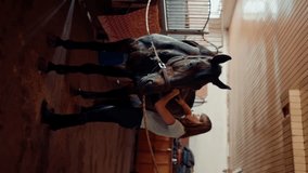 vertical video A female rider saddles her thoroughbred horse in a stable on farm preparing for equestrian training
