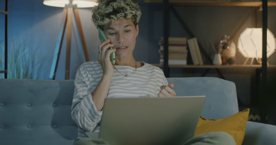 Young woman freelancer speaking on mobile phone and using laptop working from home in evening. Business communication and freelance job concept.