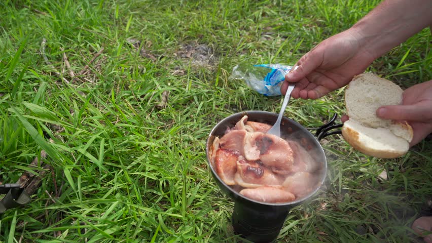 The hands of a person making a bacon sandwich while camping on grass