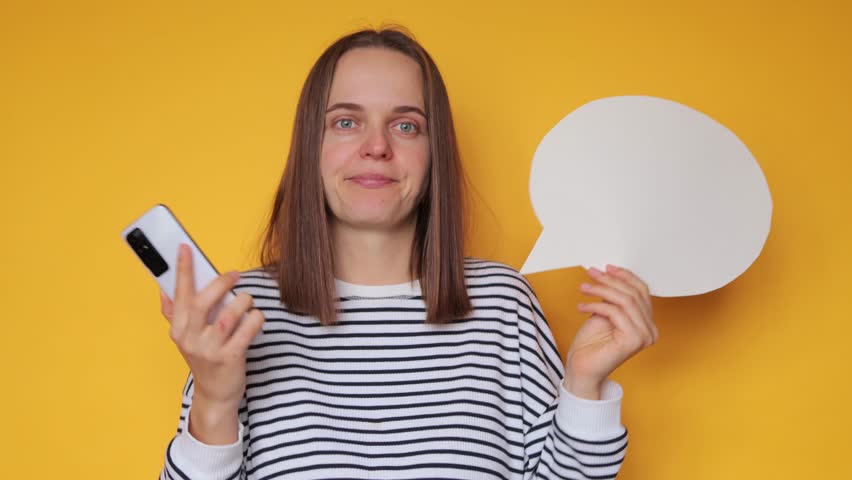 Puzzled woman wearing striped shirt posing with blank speech bubble for your promotional text against yellow wall holding smartphone shrugging shoulders doesn't know what comment to leave. Royalty-Free Stock Footage #1110374491