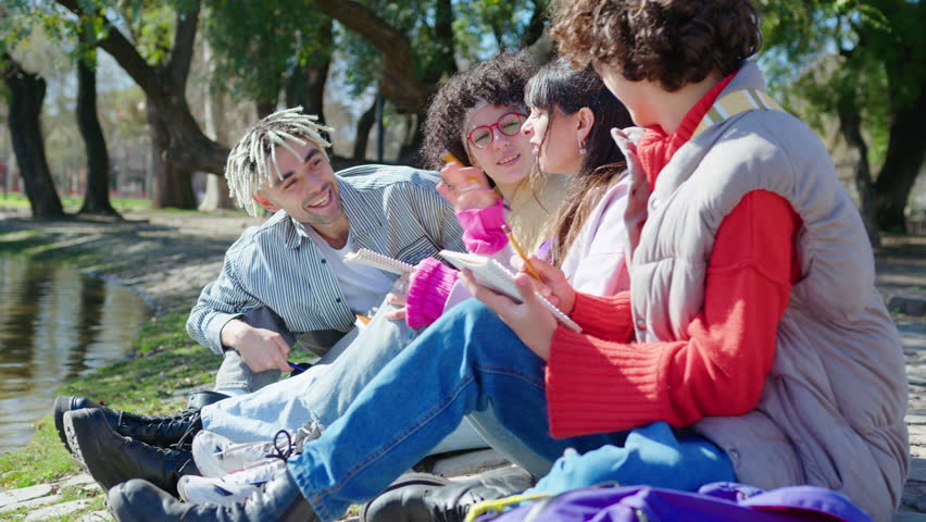 Company of young college friends sitting by pond in the park, doing homework together, smiling and chatting outdoors on sunny day