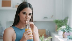 Video of beautiful sporty woman drinking healthy orange juice while standing in the kitchen at home