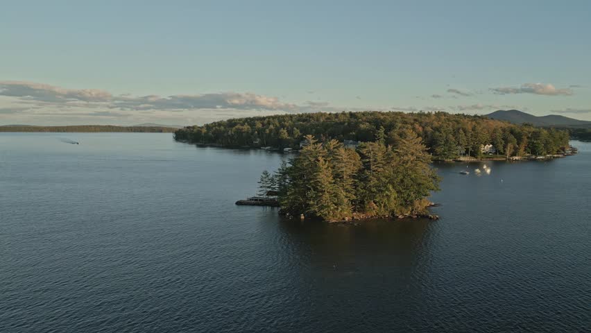 Aerial drone orbit shot captures an island nestled amidst the serene waters of Lake Winnipesaukee, USA. The island is blanketed in lush greenery, with trees create a tranquil oasis in autumn season.