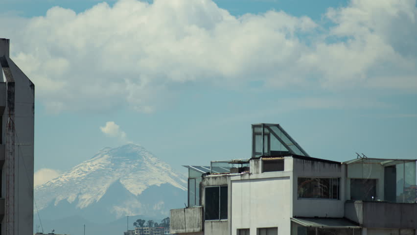 Time-lapse of view of the Cotopaxi volcano between buildings seen from Quito with a cloudy sky passing quickly against a blue sky