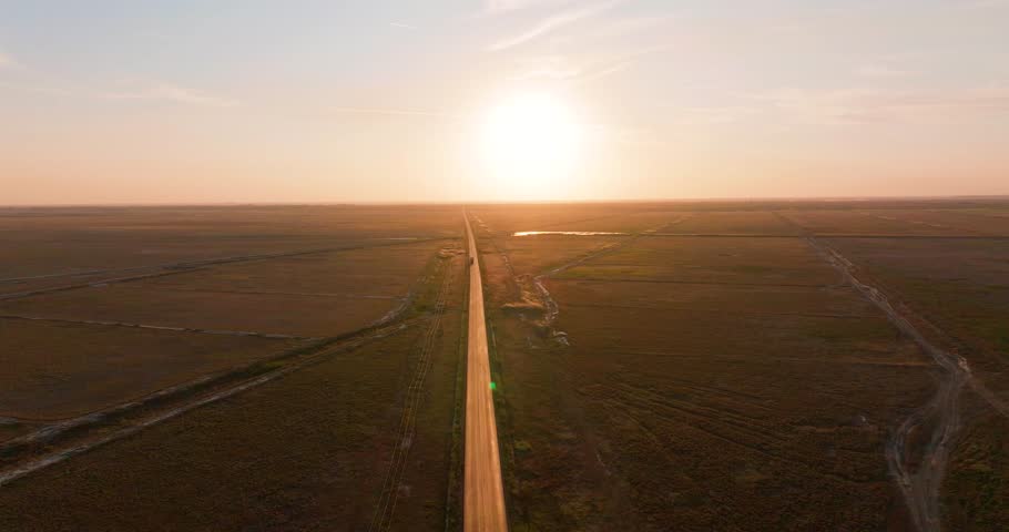 Aerial tracking shot with a transport truck that glides along a straight, sun-kissed road. The dynamic motion lend a cinematic touch to the scene. The radiant glow of the sun in the background 4K60Fps Royalty-Free Stock Footage #1110388545