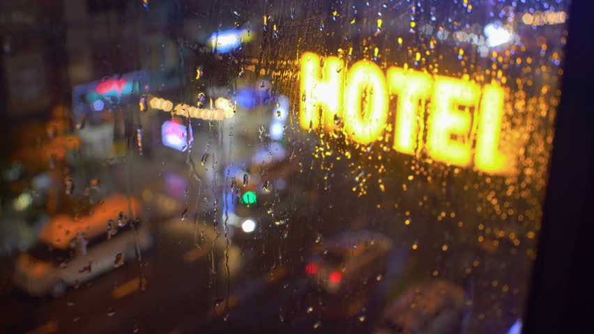 Big neon hotel sign in street below. View from building window. Rain in the city