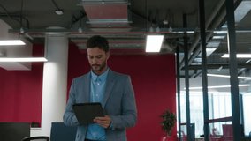 Young man holding digital tablet walking and waving at coworkers in office