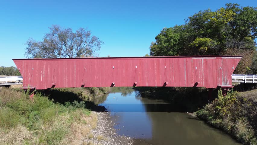 Hogback covered bridge in Madison County Iowa. Low angle aerial shot, from below the side of the bridge, rises over the top and follows the stream. Bright sunny afternoon.