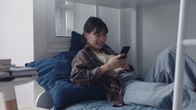 Happy young Asian Female A college student is using smartphone for chat on social network relaxing in comfortable bedroom at dormitory 