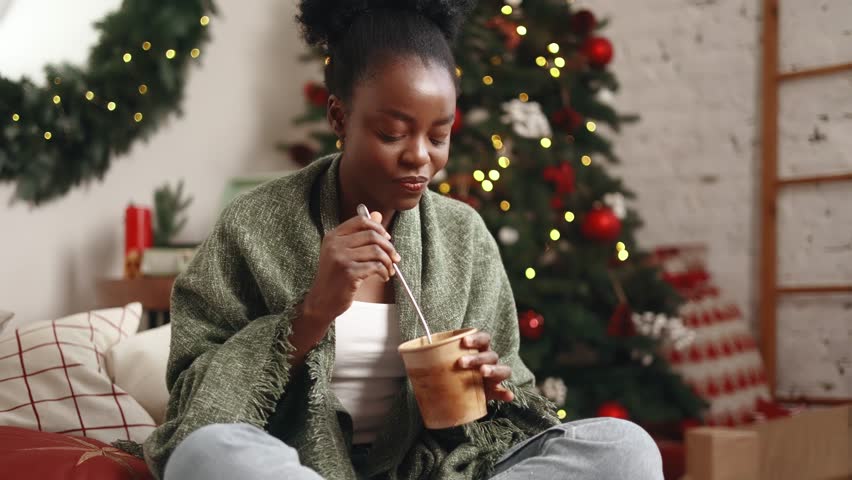 Pretty smiling young woman eating ice cream desert with great pleasure and dancing sitting under the blanket near Christmas tree at home Break the diet or tasty sweet on holidays concept