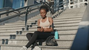 Camera view of beautiful African American female sitting on staircase outside. Watching funny video on her smartphone. Her water bottle and backpack laying next to her. Male running down in background