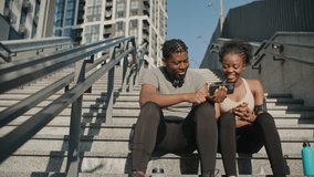 People sitting on concrete stairs somewhere in city. Two African American friends having good time together. Male showing video on his mobile device to female. Resting after exercise.
