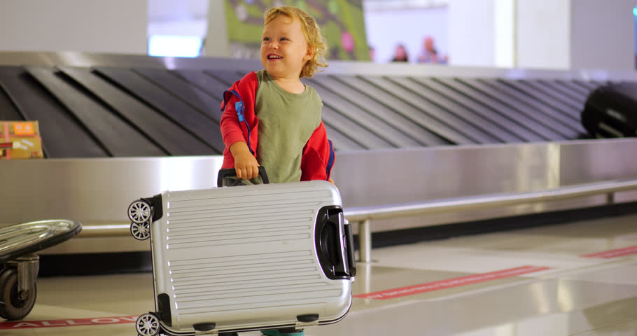Cheerful toddler plays in airport baggage claim area, lifting, dropping, and flipping small suitcase in slow motion while waiting for luggage. Selective focus, blurred conveyor belt and bags on back