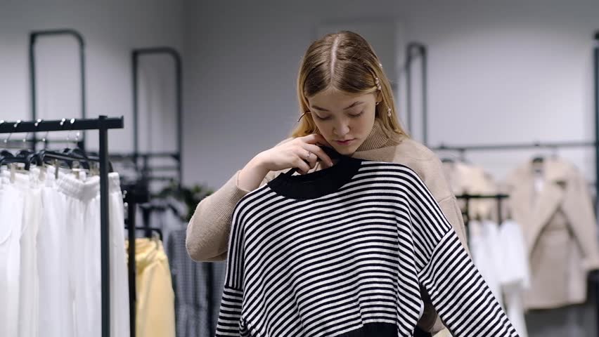 A girl tries on a striped sweater in a store. A young woman chooses clothes in a store while shopping. Seasonal holiday discounts on purchases. Black Friday Royalty-Free Stock Footage #1110426481