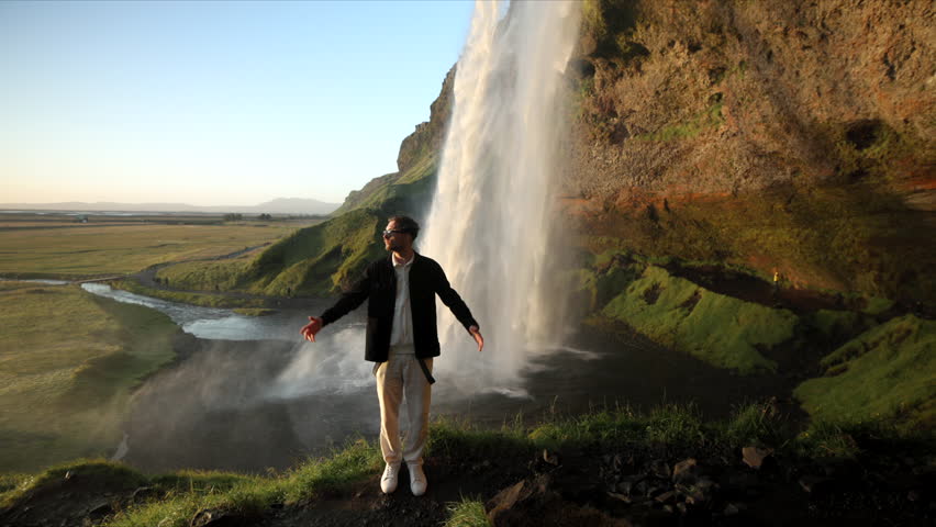 Middle-aged man in sun glasses admiring the beauty and majesty of icelandic highlands with immense waterfall dropping down creating mist behind him. High quality 4k footage Royalty-Free Stock Footage #1110429021