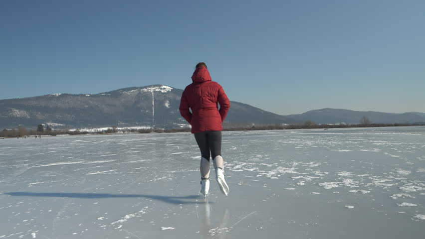 SLOW MOTION: Sporty woman ice skating across naturally frozen lake under a snowy mountain on a sunny day. A wonderful outdoor sports activity for cold winter days in the beautiful wintry countryside.