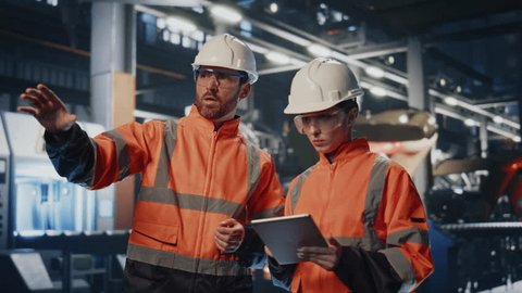 Couple industrial coworkers examining production facility in protective helmets uniform. Busy factory specialists watching machinery making notes in tablet. Professional two engineer working together : vidéo de stock
