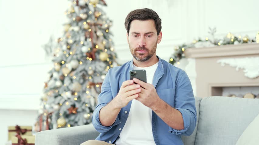 Frustrated stressed man reads bad news on smartphone sitting at home during winter New Year Christmas Xmas holidays. Worried depressed male is sad while reviewing negative message on mobile phone Royalty-Free Stock Footage #1110444933
