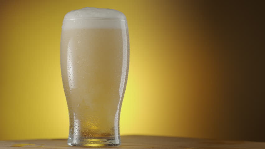 Glass of beer rotates slowly on a yellow background, there is condensation on the beer glass and there are a lot of bubbles and foam inside. A great beer background for your projects.
