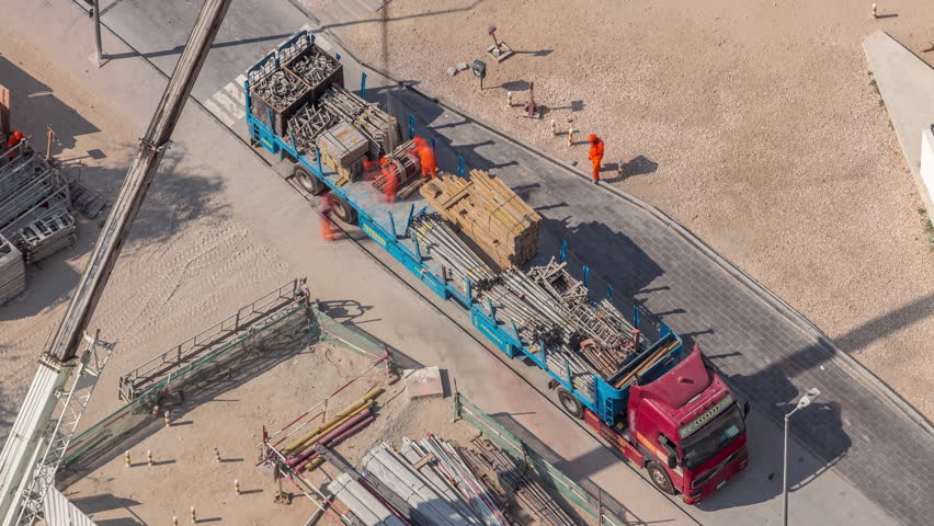 Unloading of building materials by crane at the construction site aerial timelapse. Many workers moving reinforcement. Heavy machinery